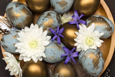 Paskalya bayramı tebrik kartı. Gri mavi altın yumurtalı Paskalya Süslemesi, bıldırcın tüyleri, koyu ahşap arka planda beyaz çiçekler. Mutlu Paskalya kartı konsepti, üst görünüm.