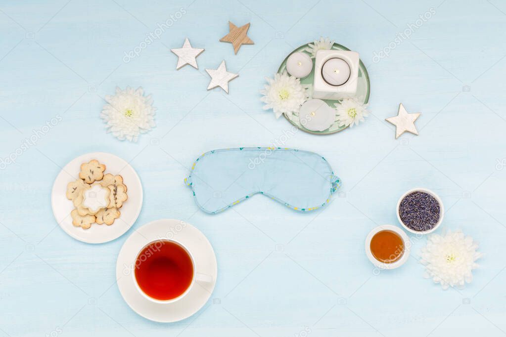 Cotton sleep mask, homemade cookies, cup of herbal tea, dry lavender flowers and candles on blue background. Top view, copy space. Healthy night sleep creative concept in blue colors. 