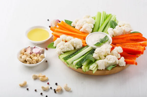 Colorful slices of raw vegetables. Carrot and cucumber sticks, pieces pepper with Vegan cashew cream sauce on white wooden background. Concept of diet, healthy and vegetarian food.