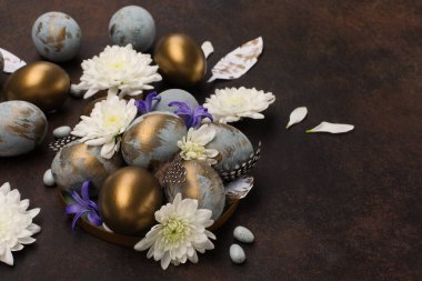 Paskalya bayramı tebrik kartı. Gri mavi altın yumurtalı Paskalya Süslemesi, bıldırcın tüyleri, koyu ahşap arka planda beyaz çiçekler. Mutlu Paskalya Sıfır Atık Konsepti, kopyalama alanı.