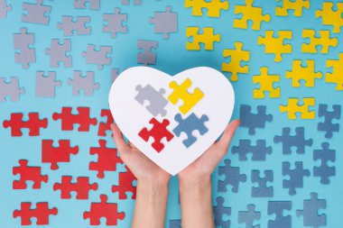 2 Nisan Otizm Dünya Farkındalık Günü için yaratıcı konsept tasarımı. Eller büyük beyaz kağıt kalp ve mavi arka planda renkli yapboz unsurları tutuyor. Üst manzara, düz uzanma.