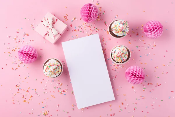 Festive holiday greeting card for Birthday, Woman or Mothers Day. Empty white blank, gift box, paper decorations and cupcakes on pink sprinkles background. Flat lay, top view, copy space.