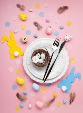 Paskalya bahar tatili sofrası. Beyaz tabak, çatal bıçak takımı, yumuşak tavşan, boyalı yumurta, açık pembe arka planda bahar çiçekleri. Şenlikli yemek sahnesi
