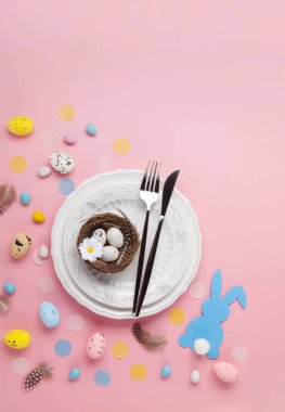 Paskalya bahar tatili sofrası. Beyaz tabak, çatal bıçak takımı, yumuşak tavşan, boyalı yumurta, açık pembe arka planda bahar çiçekleri. Şenlikli yemek sahnesi