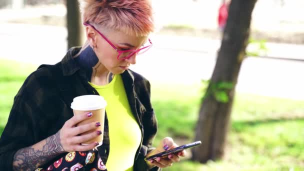 当代的嬉皮士纹身女孩在眼镜与短粉色头发与智能手机和咖啡 室外工作时间 — 图库视频影像