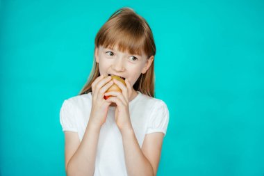 Turkuaz arka planda beyaz tişörtlü tatlı bir kız elmayı ısırıyor.
