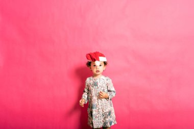 İki yaşında bir bebek, yanlış yere yerleştirilmiş Noel Baba şapkası ve komik bir görünüşü var. Pembe arka planda izole edilmiş..