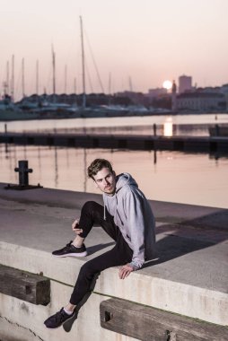 Genç kızıl saçlı adam, gün batımında gri kazak giyen bir rıhtımın yanında poz veriyor..