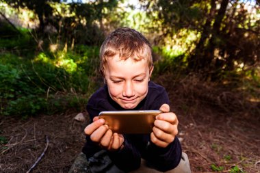 Büyük bir eğlenceye sahip bir çocuk ormanda akıllı telefonuyla video izlerken harika vakit geçirir..