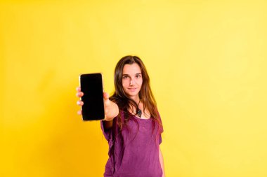 Stüdyonun sarı arka planında genç bir kadın akıllı telefonunu tutuyor..