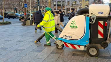 Parlak üniforma giyen bir sokak temizlikçisi Paris 'teki kaldırımı temizlemek için elektrikli süpürge kullanır. İnsanlar yakınlardaki temizlik faaliyetlerinden habersiz yürüyorlar..