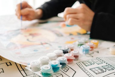 Bir insan, hafta sonu el işi seansı sırasında çeşitli renklerle çevrili basılı bir çarşafa suluboya boyayla resim yapar..