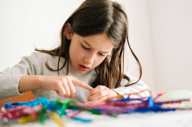 Genç bir kız evde renkli malzemeler ve malzemelerle çevrili bir tutkal tabancasıyla el sanatları faaliyetlerinde bulunuyor..