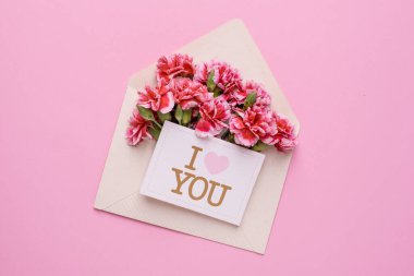 Üzerinde pembe çiçekler ve bir kart olan bir zarf. Pembe arka planda seni seviyorum.