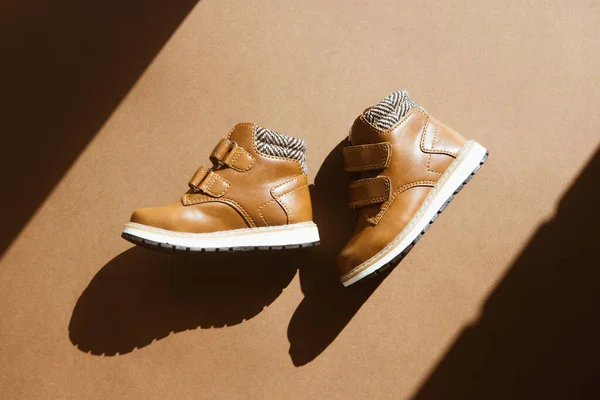 Açık kahverengi arka planda, üzerinde güneş gölgesi olan küçük bir çift deri ayakkabı. Sıcak, kürk kaplı bağcıklı çocuk ayakkabıları. Gelecek sonbahar ve kış manzarası kavramı