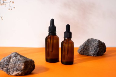Kahverengi serum ya da yağ bardağı, kurutulmuş çiçek ve ahşap formlarla turuncu arka planda pipeti olan şişeyi taklit eder. Doğal organik kozmetik, kozmetik, dermatoloji kavramı.