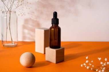 Kahverengi serum ya da yağ bardağı, kurutulmuş çiçek ve ahşap formlarla turuncu arka planda pipeti olan şişeyi taklit eder. Doğal organik kozmetik, kozmetik, dermatoloji kavramı.