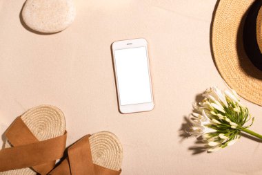 Plaj arka planı cep telefonu, kahverengi sandaletler ve güneşte kumlu bir sahilde taş. Kavram: Sıcak bir ülkede yaz tatili. 