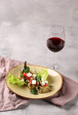 Doğu mutfağı. Bishtak patlıcan. Peynirli ızgara patlıcan, domates, zeytinyağı, sarımsak, bej bir tabakta otlar ve gri masada bir bardak kırmızı şarap. Arkaplan resmi, alanı kopyala