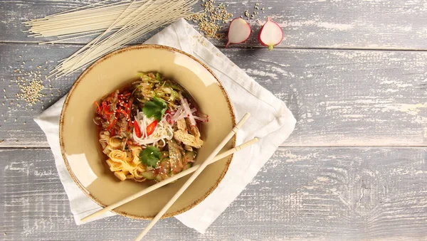 Korean cuisine. Kuksi. Korean soup with veal, wheat noodles, pickled cabbage, cucumbers, radish, omelette, asparagus and chili pepper in a bowl with sticks on wooden table. Background image, top view