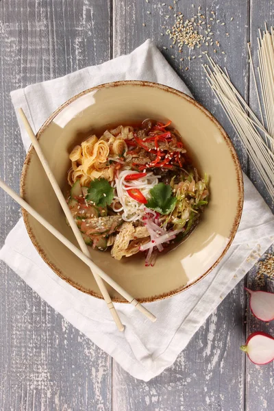 Korean cuisine. Kuksi. Korean soup with veal, wheat noodles, pickled cabbage, cucumbers, radish, omelette, asparagus and chili pepper in a bowl with sticks on wooden table. Background image, top view