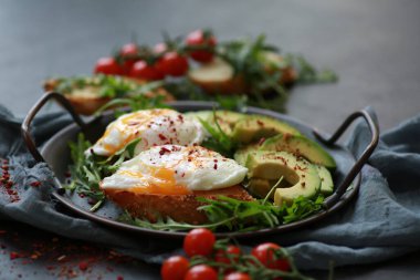 Sağlıklı kahvaltı. Kavrulmuş yumurta, roka, avokado, baharat ve kiraz domatesleri siyah bir masada gri bir tabakta. Arkaplan resmi, kopyalama alanı, yatay