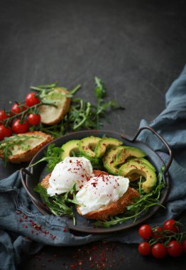 Sağlıklı kahvaltı. Kavrulmuş yumurta, roka, avokado, baharat ve kiraz domatesleri siyah bir masada gri bir tabakta. Arkaplan resmi, kopyalama alanı, dikey