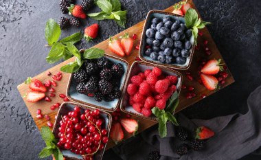 Taze meyveler. Kırmızı frenk üzümü, böğürtlen, böğürtlen ve ahududu büyük ahşap bir tahtanın üzerinde, içinde nane yaprağı, çilek ve nar tohumları var. Arkaplan resmi, üst görünüm, yatay