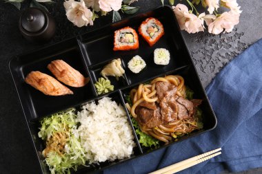 Japon mutfağı. Kara kutuda iş yemeği: pirinç, rulo, salata, siyah masada suşi. Arkaplan resmi, alanı kopyala