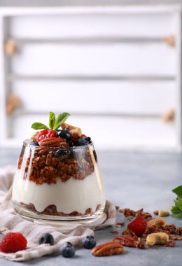 Sağlıklı kahvaltı. Yoğurt, granola, temiz bardakta ballı taze böğürtlen. Gri ışıklı arka planda fındık ve taze nane. Arkaplan resmi, kopyalama alanı, dikey