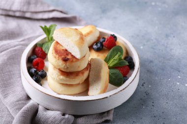 Sağlıklı kahvaltı. Peynirli kekler, ballı süzme peynirli krepler, taze böğürtlenler, gri masadaki beyaz ahşap bir tabakta nane şekeri. Arkaplan resmi, kopyalama alanı, yatay