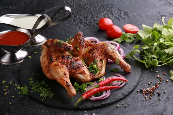 Fried chicken with fresh herbs, coriander, vegetables, onions, spices and sauces on a black table. Tobacco chicken. Background image, copy space