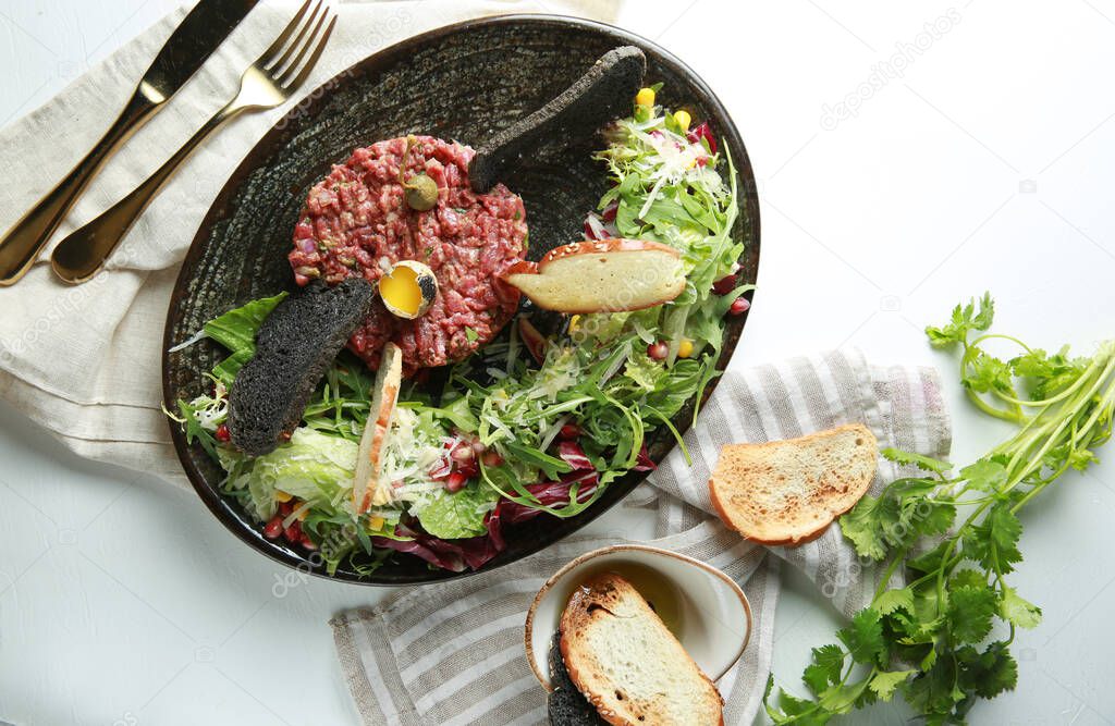 Tartare with crouton with herbs on a dark plate with egg and spices on white table, cilantro, knife, fork. Restaurant menu. Background image, copy space