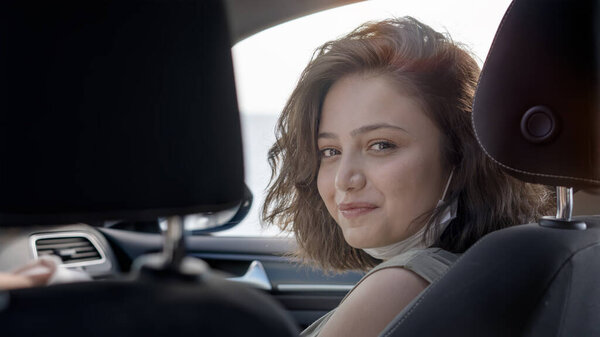 Young beautiful woman in mask sitting on the front passenger seat inside of a car smiles looking over shoulder at camera.