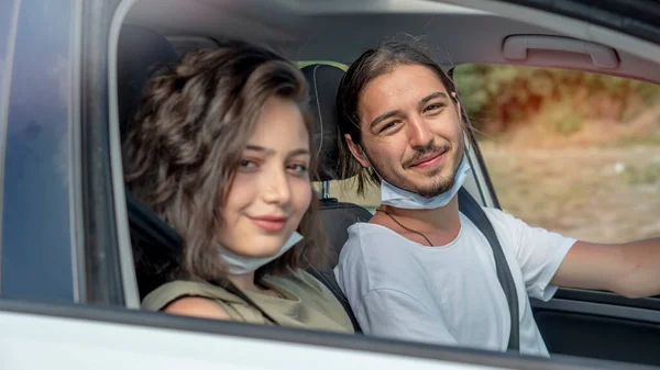 Happy young couple, friends with face masks sitting in the car and talking each other. Road trip in new normality. Corona virus pandemic concept.