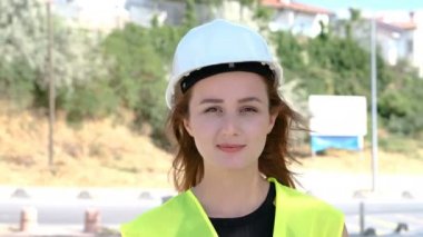 Üniformalı, koruyucu beyaz kasklı, gülümseyen ve inşaat alanında kameraya bakan çekici bir kadın mühendis ya da işçi portresi. 4k Çözünürlük, yavaş çekim