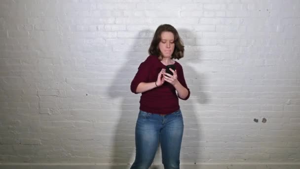 Een jonge vrouw dansen speels voor een witte bakstenen muur met haar mobiele telefoon. — Stockvideo