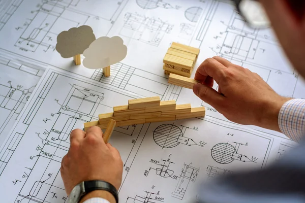 Arsitek Man menggambar rencana, grafik, desain, bentuk geometris dengan pensil pada lembaran besar kertas di meja kantor dan membangun model rumah dari balok kayu (bar ) Stok Foto Bebas Royalti