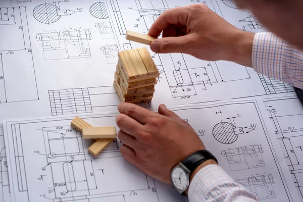 Arsitek Man menggambar rencana, grafik, desain, bentuk geometris dengan pensil pada lembaran besar kertas di meja kantor dan membangun model rumah dari balok kayu (bar ) Stok Lukisan  