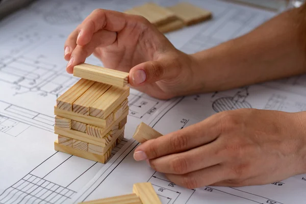 Man architect draws a plan, graph, design, geometric shapes by pencil on large sheet of paper at office desk and builds model house from wooden blocks (bars) — Stock Photo, Image
