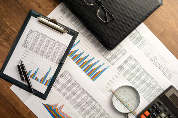 Business accessories (notebook, calculator, planchette, tablet, fountain pen, glasses) and graphics, tables, charts on white sheets on office desk. Soft focus.