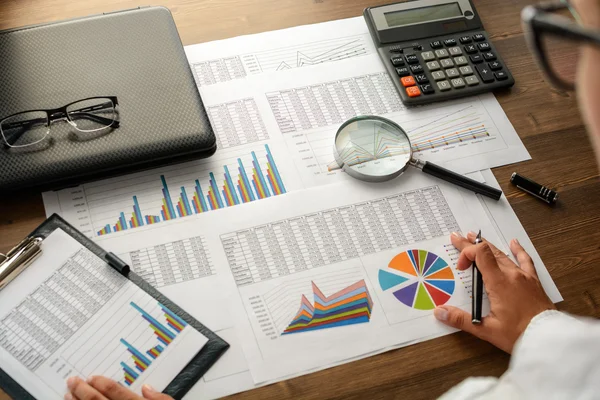 Girl and business accessories (notebook, calculator, planchette, tablet, fountain pen, glasses) and graphics, tables, charts on white sheets on office desk. Soft focus.