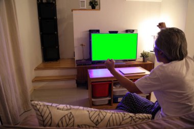 Kanepede oturan kıdemli Asyalı adam televizyon izliyor, kamera televizyona odaklanıyor..