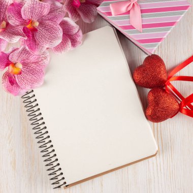 Sevgililer Günü arkaplanı boş not defteri, hediye kutusu, pembe orkide çiçeği ve beyaz tahta yüzeyde kırmızı kalplerle dolu. Düz yatış, üst görünüm.
