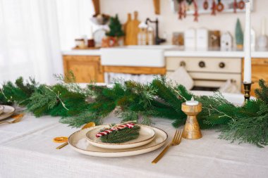 Noel dekoru. Güzel düzenlenmiş yemek masası, zarif beyaz tabaklar, beyaz masa örtüsü üzerinde altın çatal bıçak takımı ve ladin çelengi, aile ve arkadaşlarla yemek için bayram atmosferi yaratıyor..