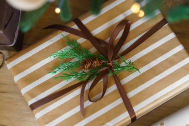 Paketlenmiş Noel hediyesi, çizgili kağıt ve kahverengi kurdeleli hediye kutusu, Noel ağacının altında ahşap yüzeyde çam kozalağı ve yeşil dal süslemesi, sıcak ve şenlikli kış tatili havasını yakalıyor.. 