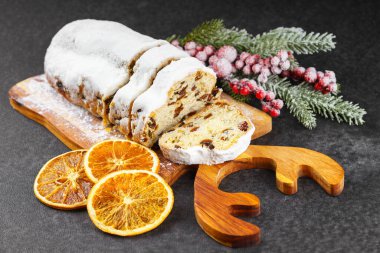 Geleneksel Noel süslemesi, ahşap tahtada dilimlenmiş tatlı meyve somunu, siyah desenli arka planda toz şekeri. Baharatlar, kurutulmuş meyveler ve şenlikli dekorasyonlar kış tatili havası yaratır.