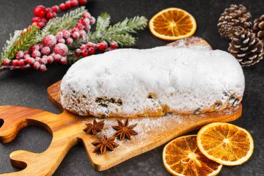 Klasik Noel Stollen, ahşap tahtada kurutulmuş portakallar, siyah desenli yüzey üzerinde çam dalları ve koniler ile pudra şekerli kremayla kaplıdır. Geleneksel bayram yemeği, tatlı meyveli ekmek.