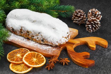 Klasik Noel Stollen, ahşap tahtada kurutulmuş portakallar, siyah desenli yüzey üzerinde çam dalları ve koniler ile pudra şekerli kremayla kaplıdır. Geleneksel bayram yemeği, tatlı meyveli ekmek.