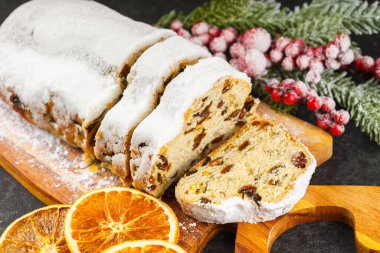 Geleneksel Noel süslemesi, ahşap tahtada dilimlenmiş tatlı meyve somunu, siyah desenli arka planda toz şekeri. Baharatlar, kurutulmuş meyveler ve şenlikli dekorasyonlar kış tatili havası yaratır.